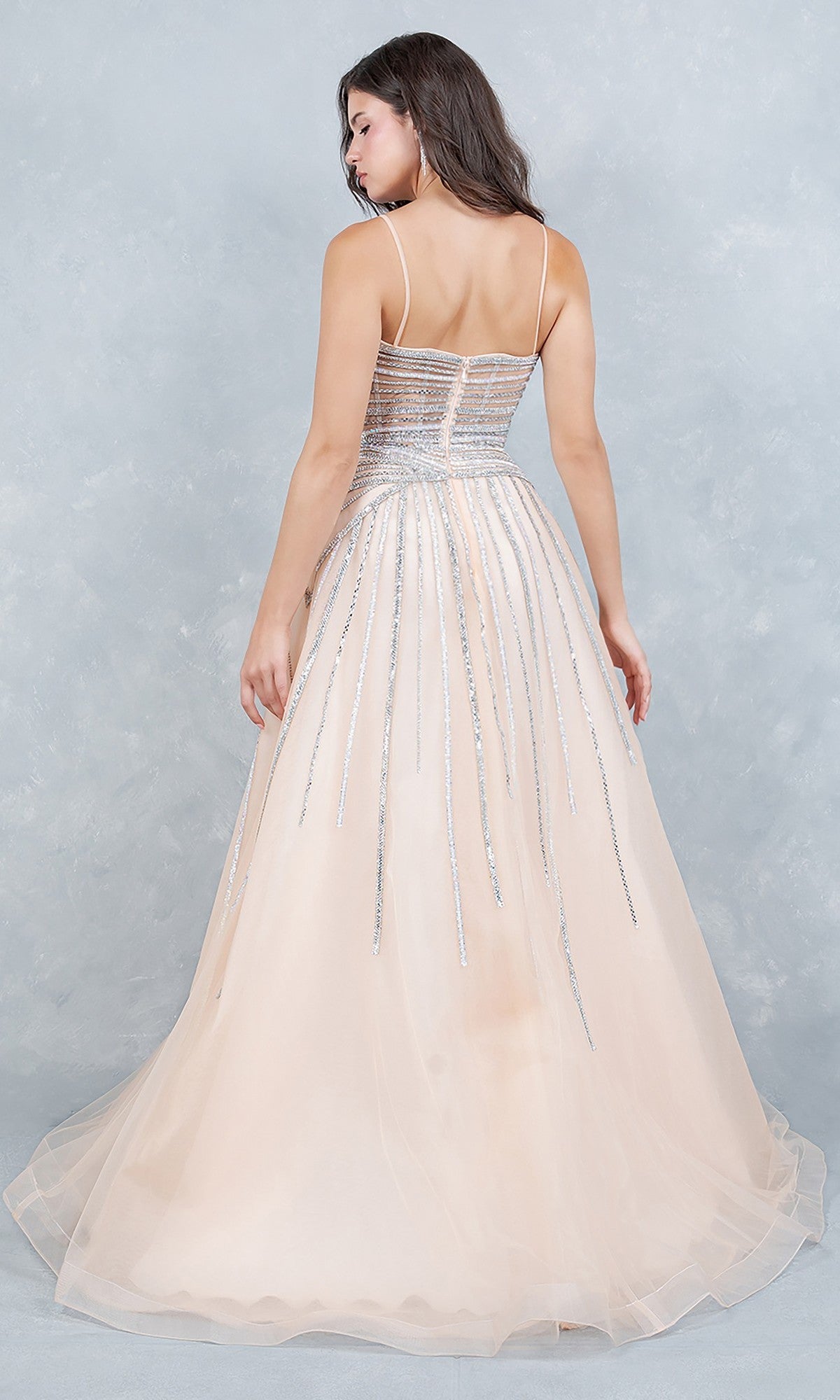 A woman stands with her back to the camera in a light peach, floor-length Cinderella Couture 8569J prom dress featuring thin straps, beaded and silver sequin detailing, set against a light textured background.